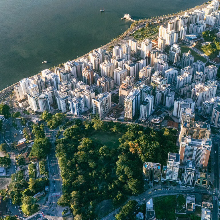 An expansive aerial view of a dense coastal city featuring high-rise buildings, residential skyscrapers, a lush green park forest, and a busy seaside highway running alongside the vast blue ocean bay during late afternoon sunlight.